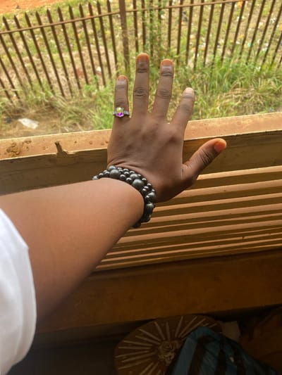 The image captures a close-up perspective of a person's right arm and hand, extending outwards from what appears to be an interior space towards an outdoor view. The arm and hand have a dark skin tone.
The hand is adorned with two distinct accessories. On the ring finger, a silver-toned ring features a prominent, oval-shaped gemstone with a striking iridescent quality, displaying hues of purple, green, and yellow, surrounded by smaller, clear stones. Around the wrist, a bracelet composed of multiple strands of dark, spherical beads, likely black or dark grey, adds texture and style. A portion of a white sleeve is visible at the bottom right, suggesting the person is wearing a white garment.
The hand is positioned near or resting on a sturdy, possibly metallic, vertical structure that forms part of a window frame or railing. Beyond this structure, a series of parallel vertical bars are visible, casting distinct shadows on adjacent surfaces, indicating bright sunlight. Through these bars, an outdoor scene unfolds, characterized by vibrant green grass, interspersed with what appears to be scattered litter or debris. The overall impression of the outdoor area is somewhat unkempt, yet bathed in natural light.
To the right side of the image, closer to the interior, parts of another structure or wall are visible, rendered in darker, earthier tones due to less direct light. In the upper right corner, indistinct objects are partially visible; one appears to be a basket-like item with radiating patterns, and another is a folded blue fabric or bag.
The lighting suggests it is daytime, likely a sunny day, given the sharp shadows cast by the bars. The photograph is taken from a high angle, looking down and out, creating a contemplative or observational mood. There is no visible text in the image.