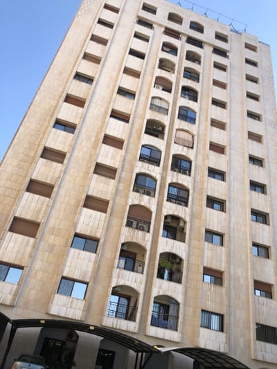 The image displays a multi-story residential building located in Kafr Sousa Municipality, Syria, viewed from a low, upward angle against a clear blue sky. The building features a light-colored, possibly limestone or tiled, facade with pronounced vertical columns separating recessed sections containing windows and balconies.
The structure appears to have at least 12 visible stories. Each story features a repeating pattern of rectangular windows and arched balconies. Many windows are fitted with light brown external roller shutters, while others are glass, reflecting the blue sky. The balconies, distinguished by dark metal railings, frequently house external air conditioning units. On one mid-level balcony, a green potted plant or small tree is visible, and on another lower balcony, light-colored laundry or textiles are hanging.
At the base of the building, a dark, arched carport or shade structure extends, underneath which a light-colored vehicle, possibly silver or light blue, is partially visible. The overall environment suggests a well-maintained urban residential area under bright daylight. No individuals are present in the scene.