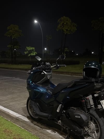 The image, taken at night in Bekasi Regency, Indonesia, prominently features a dark teal or blue Honda ADV 160 scooter. A dark green full-face motorcycle helmet rests on the rear of the scooter's seat.

The scooter is parked on a paved surface, likely a road or parking lot, which is partially delineated by a white line. Adjacent to the pavement is a strip of green grass, followed by several tall trees with visible foliage, silhouetted against the dark night sky. The scene is illuminated by multiple streetlights, whose bright glow pierces the darkness, highlighting elements of the foreground and casting ambient light on the background.

No people are present in the image, contributing to a quiet and solitary atmosphere. Small water droplets or dust particles are visible on the helmet and various surfaces of the scooter. A portion of a license plate or tag, displaying the letter 'B', is visible on the rear fender of the scooter. The Honda logo is also visible on the engine casing. The overall setting suggests an urban or suburban roadside or park area after dark.