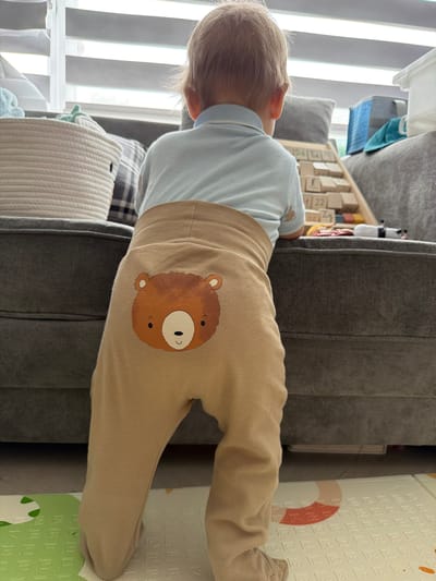 A young child with blonde hair, seen from behind and from the side, is leaning over a grey sofa. The child is wearing a light blue t-shirt and tan sweatpants with a graphic of a teddy bear's face printed on the back of the right leg. The teddy bear graphic is brown with a white face, black eyes, and a light brown nose. The child's head is tilted downwards, and their hair is short and wispy.
The background includes a wooden toy shelf filled with blocks, some of which have letters and numbers on them. One block is visible with the number "22" on it. To the right of the sofa, a portion of a white mat with embossed letters and numbers can be seen, along with a curved red and white design. The setting appears to be indoors, possibly a living room or play area, with blinds visible in the background suggesting natural light from a window. The overall impression is of a candid moment, possibly during playtime. There is no discernible text related to a specific brand or message.