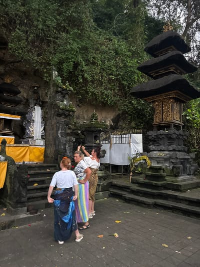 In Dawan, Indonesia, three women are gathered in front of a stone temple structure. The temple features multiple tiers topped with black thatched roofs and ornate golden decorations. To the left, another temple structure is visible with yellow fabric draped around its base. The ground is paved with dark stone tiles, and fallen leaves are scattered across it. Lush green foliage covers the rocky hillside behind the temples. One woman, on the right, is taking a photo with her phone. Another woman in the center is looking towards the temple, while the woman on the left is adjusting her sarong. The lighting suggests it is late afternoon or early evening.