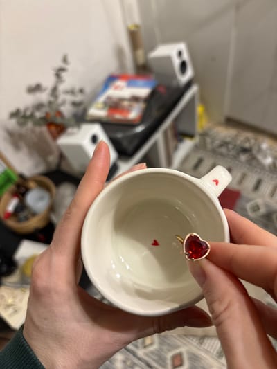 The scene is set indoors in Kudrovo, Russia. Two human hands, appearing to belong to a single individual, are prominently featured in the foreground. The left hand grasps the lower body of a white ceramic mug, while the right hand holds a gold-toned ring with a large, heart-shaped red gemstone, presenting it towards the mug's interior. Inside the mug, at its base, a single small red heart-shaped decal or piece of confetti is visible. A similar small red heart decal is present on the mug's handle. The hands display natural skin tones and visible fingernails.

The background, largely out of focus, reveals an interior room. A light-colored wall is visible behind a shelf unit. On the shelf, two white cube-shaped speakers are placed. Other indistinct objects include a magazine or book with a red element on its cover, a small potted plant with sparse foliage, and a wicker basket containing various items. A patterned rug is partially visible on the floor to the right. The lighting suggests an indoor artificial source.
