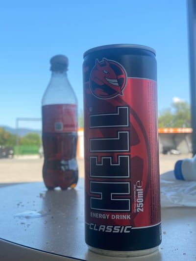 An outdoor scene with a blue sky dominates the background. In the foreground, a red and black can of "HELL ENERGY DRINK CLASSIC" stands upright on a speckled white surface, possibly a table. The can is partially obscured by its own label, which prominently features the "HELL" logo with a red devil icon. To the left and slightly out of focus is a plastic bottle filled with a dark brown liquid, likely a beverage. The surface also shows scattered crumbs and a small puddle, suggesting a recent consumption or spillage. The background is blurred but hints at an urban or semi-urban environment with some structures and greenery. The setting appears to be the city of Kran, Bulgaria.