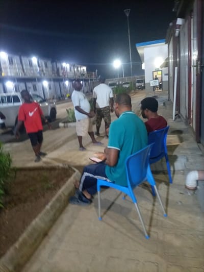 At night, several people gather outside near some buildings. A man in a green shirt sits on a blue chair, holding something. Another man sits on a similar chair behind him. A man in a red shirt walks nearby, while three others stand and talk. The area is lit by streetlights and lights from the buildings. A white pickup truck is parked in the background.