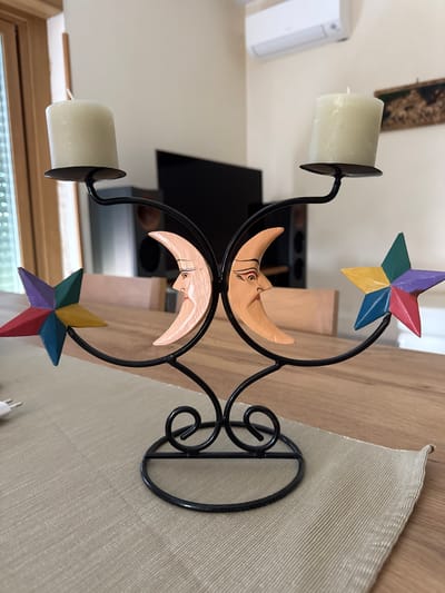 The image depicts a decorative candle holder with two candles. The candle holder is made of black metal and features two crescent moons and two stars. The moons are wooden with carved faces, and the stars are painted in multiple colors, including red, blue, yellow, green, and purple. The candle holder is sitting on a light-colored wooden table with a beige fabric runner. In the background, there is a window, a television, and part of a wall. There is no evidence to suggest that this image was taken in Aprilia, Italy.