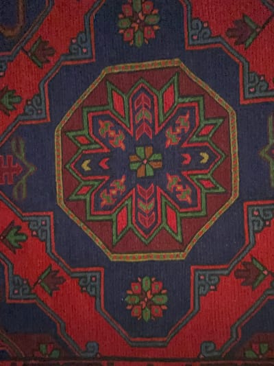 The image shows a close-up view of an intricately patterned rug. The rug features a central octagonal motif with radiating geometric shapes in shades of red, green, and dark blue. The outer border is a deep red, interspersed with dark blue geometric elements and floral designs. The texture of the rug, with its visible weave, suggests it is likely made of wool. The lighting is even, highlighting the colors and patterns without harsh shadows. No specific location in Saint-Quentin, France, can be identified from the image alone, as it focuses solely on the textile.