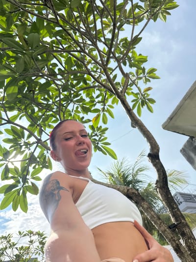 A female individual is captured in an upward-angled close-up against a backdrop of tropical foliage and a bright sky in Kerobokan, Indonesia. Her reddish hair is pulled back, and she has a hoop piercing in her left nostril and a barbell piercing in her lower lip. Her eyes are partially closed, and her tongue is extended. She wears a white crop top. A dark, avian-themed tattoo is visible on her upper left arm. The immediate environment includes dense green leaves and branches of a large tree. In the background, sections of light-colored building structures and additional palm foliage are visible under a blue sky with scattered white clouds.