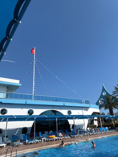 This media file depicts a vibrant outdoor scene at a resort, likely during daytime given the bright sunlight and clear blue sky. The primary subjects include people enjoying a swimming pool and lounging chairs set up around it. The scene is dominated by a large, multi-story building with a striking blue and white facade, designed to resemble a ship. This architectural feature is a prominent object in the frame.
Several people are visible in and around the pool. In the water, two individuals appear to be playing with a soccer ball and a goal net, suggesting a recreational activity. Another person is partially submerged in the pool. Around the pool, numerous blue and white lounge chairs are arranged, some occupied by people relaxing. Various objects like towels and a yellow inflatable ring are also present.
A notable detail is a string of small, triangular flags, alternating between white and blue, stretched across the sky, adding a festive atmosphere. A red flag, possibly a national flag, is also flying from a tall mast. Palm trees are visible in the background, hinting at a warm, tropical or subtropical climate.
The overall emotion conveyed is one of leisure and enjoyment, characteristic of a holiday setting. The location is suggested to be Manavgat, Turkey, which aligns with the presence of palm trees and the sunny, clear weather. No visible text is present in the image.