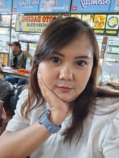 A woman with long brown hair and a white shirt is shown in a close-up shot. She rests her face on her hand, wearing a gray watch. The background reveals a food stand with a menu board advertising "SOTO DAGING" or meat stew. There is another person wearing a cap and jacket seated in the background, possibly at a table in the eatery.