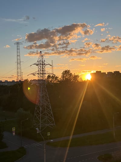 This image depicts a sunset scene in Shushary, Russia. The sun, low on the horizon, casts a warm, golden glow across the sky, illuminating scattered, wispy clouds. The light creates strong sunbeams that stretch across the frame, contributing to the dramatic atmosphere.

Dominating the midground are two large electricity pylons, their intricate metal latticework silhouetted against the bright sky. The taller pylon stands closer to the viewer, while the second is slightly further away. Trees line the background, their foliage dense and dark, contrasting with the illuminated sky. In the distance, the outlines of buildings are visible, suggesting an urban or suburban setting.

The foreground features a road, likely a street or highway, with clear lane markings. Several traffic signs are visible along the roadside, including pedestrian crossing signs and what appear to be speed limit signs. The overall scene evokes a sense of the end of the day, with the natural beauty of the sunset juxtaposed with man-made infrastructure. The lighting suggests it is late afternoon or early evening. There is no visible text in the image other than on the traffic signs.