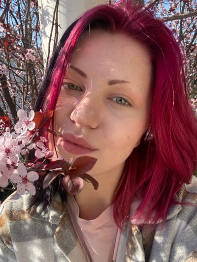 The image displays a close-up, outdoor portrait of an individual in Kunje, Montenegro. The subject is an adult female with fair skin, green eyes, and prominent pink-magenta hair, featuring a darker, possibly black or dark brown, streak on the left side. Her eyebrows are light brown and arched. Her lips are full and light pink. She wears a light-colored, possibly plaid or checkered, collared jacket over a light pink t-shirt. A metallic earring is visible on her right ear. A branch featuring light pink blossoms and dark red leaves is positioned diagonally across the lower left foreground, extending towards the subject's face and shoulder. Bright sunlight illuminates the scene, casting noticeable shadows on the subject's face and upper body. The background is blurred, showing a tree with similar light pink blossoms against a clear blue sky, with a light-colored architectural element partially visible in the upper left. The individual is looking directly towards the camera.