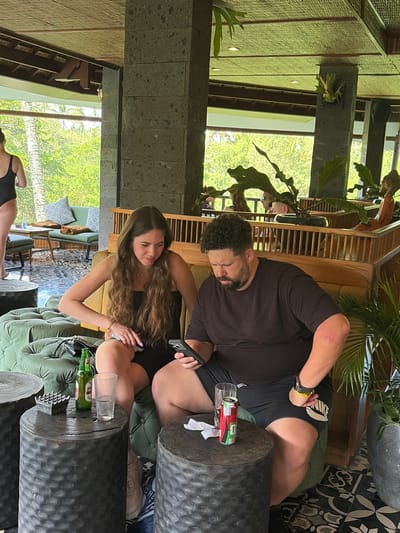 In Tegallalang, Indonesia, a woman with long brown hair and a black dress sits on a green tufted couch, looking at her phone. Next to her, a man with curly dark hair and a black t-shirt and shorts is also looking at his phone. They are seated in a lounge area with dark, textured cylindrical tables. On one table, there is a green bottle of beer and a glass of water, along with an ashtray. On another table, a can of Coca-Cola and a piece of white paper are visible. In the background, a woman in a black swimsuit stands near a sofa and a table with cushioned seating. The environment appears to be an open-air establishment with natural light filtering through lush green foliage and trees. The architecture features dark stone columns and a thatched ceiling.
