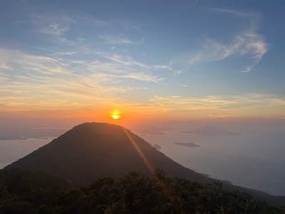 The image depicts a serene sunrise over a mountainous landscape near El Cacao, El Salvador. In the foreground, lush green foliage, likely trees and bushes, is silhouetted against the bright sky. A prominent, dark-hued mountain dominates the mid-ground, its slopes covered in vegetation. The sun, a brilliant orb of light, is positioned just above the mountain's peak, casting a warm orange and yellow glow across the horizon. A visible sunbeam streaks diagonally downwards from the sun. Beyond the mountain, a vast body of water, possibly a lake or ocean, is visible, dotted with several islands of varying sizes. The water appears calm, reflecting the soft colors of the sky. The sky itself transitions from a deep blue at the top to lighter shades of blue, orange, and yellow near the horizon, with wispy clouds scattered throughout. The overall scene conveys a sense of tranquility and natural beauty.
