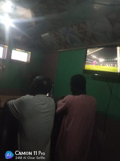 Inside a dimly lit room, two individuals are seated. One wears a white shirt and the other a pink one. Two screens display betting odds or market data on the left. A larger television on the right shows a soccer game. The room has green walls and a patterned ceiling. The "CAMON 11 Pro" watermark is visible in the lower left corner.