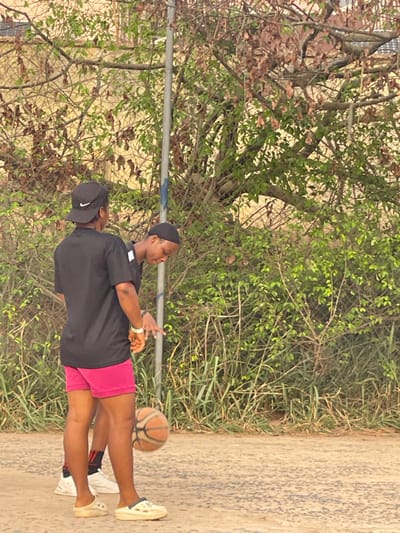 Two people stand on a concrete surface near greenery, including a tree. One person, wearing a black t-shirt, pink shorts, and light-colored sneakers, bounces a basketball. Another person stands close by wearing a black cap, a black t-shirt, and dark socks. A long, thin, horizontal pole stretches across the image behind the two people.