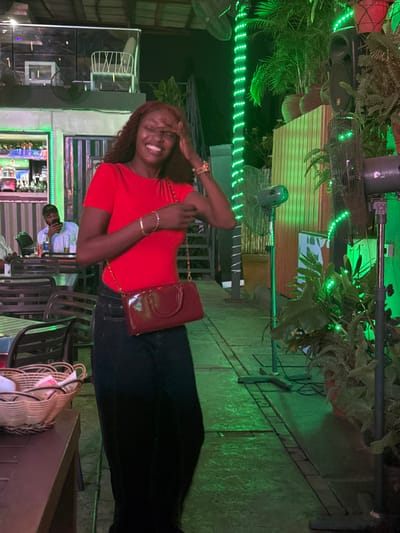 The image depicts an outdoor evening scene at a commercial establishment in Falomo, Nigeria, likely a restaurant or bar. The primary subject is a female with dark, wavy hair, positioned slightly left of center, facing the camera. She is captured with her eyes closed, mouth open in a wide smile, and her right hand touching her forehead. She wears a red short-sleeved top and dark trousers, with a glossy, reddish-brown shoulder bag featuring a gold chain strap across her body. Her left wrist displays multiple gold-colored bangles, and her right wrist has a multi-colored beaded bracelet.

In the mid-ground to the left, a male individual wearing a light-colored shirt and a dark baseball cap is seated at a table, looking down at a mobile phone. A beverage bottle is visible on his table. To the immediate foreground left, a light brown woven basket, possibly containing napkins or bread, rests on a dark brown rectangular table. Dark-colored chairs are partially visible around this table.

The background elements include a bar area on the left, displaying various bottles and glasses, illuminated by green light. Above the bar, a structure, possibly an upper seating area or balcony, features white chairs. The right side of the scene is characterized by abundant green foliage from potted plants. Several industrial-style standing fans are visible, illuminated by green light. Vertical structures, possibly poles or decorative panels, are wrapped with green LED string lights. A large black speaker is mounted in the upper right. The ground is paved with what appears to be concrete slabs or tiles, reflecting the predominant green lighting.