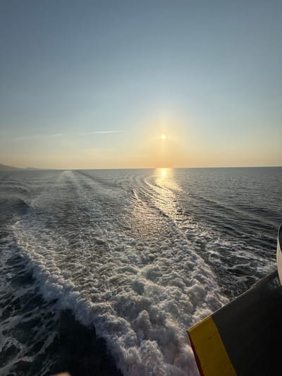 The image captures a maritime scene from the stern of a moving vessel in the waters off Kato Karioutes, Greece, during a low sun event. The sun, a bright golden orb, is centered horizontally in the upper-mid section of the frame, casting a direct, shimmering reflection path across the water towards the camera's position. The turbulent wake of the vessel, characterized by white foam and churned water, originates from the bottom-center and extends diagonally towards the mid-left of the frame. A portion of the vessel's dark grey stern, marked with a distinctive yellow and red stripe, is visible in the bottom-right corner. A faint, dark silhouette of land is discernible on the distant horizon line to the left. The sky exhibits a gradient from clear light blue at the top to a warmer, lighter hue near the horizon where the sun is situated. No individuals are present in the scene.