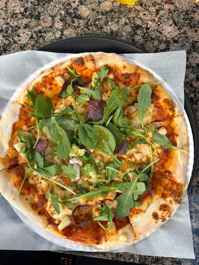 An overhead shot shows a pizza served on a white paper liner on a black tray. The pizza has a golden-brown crust and is topped with red sauce, melted cheese, and pieces of chicken. A generous portion of arugula and some red leaf lettuce are scattered on top of the pizza, glistening with dressing. The pizza is cut into slices and appears to be freshly prepared. The background is a mottled brown and black speckled countertop, possibly granite. The image is sharp and well-lit, highlighting the textures and colors of the food. The scene is likely from an indoor setting, given the lighting and countertop surface. This image was taken in Rūdiškės, Lithuania.