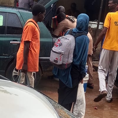 The image, taken in what appears to be a public outdoor space in Dadi Riba, Nigeria, features several individuals engaged in different activities. One man wears a yellow t-shirt with "RT KADE" printed on it and white pants with stripes down the sides. He is walking away from the camera. Another individual in a blue hooded top and black bottoms is wearing a black and white striped backpack. A man in an orange t-shirt stands beside a green vehicle. Other individuals are also visible, their clothing varying in color. The scene is crowded, with a white vehicle partially visible in the foreground.