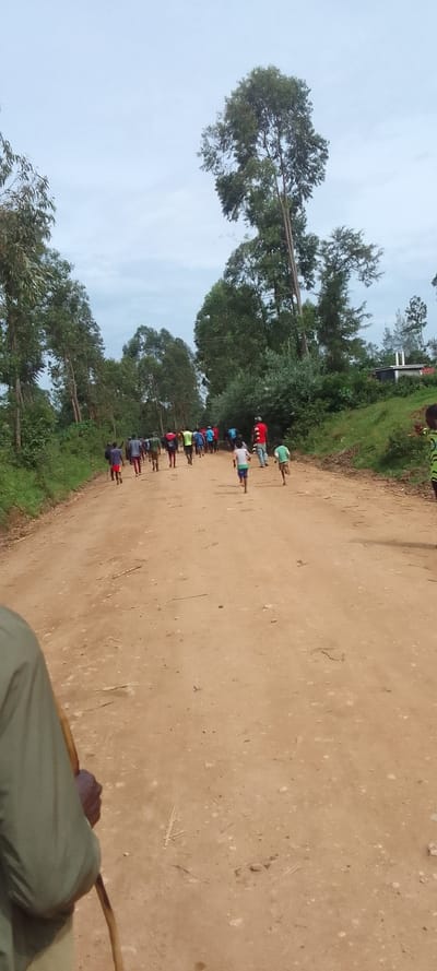 The image shows a dirt road lined with trees and bushes, with a group of people walking away from the camera, some in athletic wear. There are some people, possibly children, running towards the horizon. The location is a rural area in Lubao, Kenya, near latitude 0.384 and longitude 34.822. The scene appears to be a path or road used by people on foot. The sky is overcast with a light gray color. The photographer is holding a wooden stick and their body is partially visible in the bottom left corner.