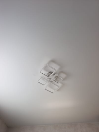 This is a photograph of a modern ceiling light fixture with four square frames arranged in a cluster, mounted on a white ceiling. The fixture is centrally located in the frame, with the majority of the image showing the plain white ceiling. The lighting creates soft shadows, highlighting the dimensionality of the fixture. The bottom of the frame shows a glimpse of a tiled wall with a horizontal trim line. The photograph appears to be taken from below, looking upwards.