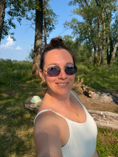 A woman with her hair in a bun, wearing sunglasses and a white top, takes a selfie outdoors. She is smiling and has a necklace on. The background shows trees, grass, and a bright blue sky. There are logs and what appears to be a fire pit in the background. The scene is likely in Ol'khovskoye, Russia, given the natural, wooded environment.