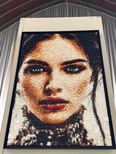 The main subject is a large, framed three-dimensional artwork, shown tilted in the image, depicting a detailed, pixelated portrait of a person's face, likely a woman. The face is composed of countless small, square and rectangular blocks of varying heights and shades, creating a mosaic-like image with a sculptural texture. Features include a prominent eye with a dark pupil and lighter iris, part of a nose, and distinctly rendered lips in reddish tones. The skin is depicted using a range of cream, beige, and brown blocks, while darker blocks form areas like hair or shadows. In the upper right corner of the actual artwork (when viewed upright), a small, circular logo or monogram resembling an 'N' is faintly visible within the white blocks.
The artwork is set within a slim black inner frame, which is then surrounded by a wider, light-colored (off-white) outer frame or mounting board. The entire framed piece is displayed indoors, likely in a gallery or exhibition space. The background, which appears vertical despite the tilt of the artwork in the photograph, shows a dark brown surface with subtle green stripes on one side and a light grey, subtly textured or pleated surface on the other. A thin, reflective metallic strip is present along the bottom edge of the outer frame. The lighting appears even, highlighting the three-dimensional nature of the blocks. No specific activity or event is depicted other than the display of the art.