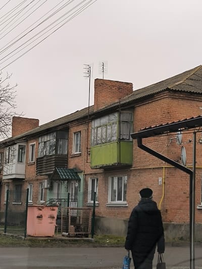 The image presents an overcast daytime scene of a street in Illintsi, Ukraine, featuring a multi-story brick residential building. The building, likely an apartment block, has a weathered grey roof and several enclosed balconies/loggias, each with a distinct appearance; one on the right is painted bright green, another in the middle has dark corrugated siding, and one on the left is white. Multiple windows are visible across the facade. Two Yagi-style television antennas are mounted on the rooftop, above brick chimney structures.
In the foreground, a person is seen from behind, walking away from the viewer on a paved surface next to the building. The individual is dressed in a dark, possibly padded, winter coat and a dark hat, carrying a blue-patterned bag in one hand and a dark bag in the other.
Notable details include overhead utility lines against the grey sky on the left. An entrance to the building features a small green arched awning and a dark metal gate. In front of this, there is a large, faded pinkish-orange dumpster-style trash bin and a metal grate enclosure, likely for refuse collection. Green metal posts mark a boundary. On the brick wall to the right, the number "10" is affixed, possibly a building or entrance number. Two white satellite dishes are visible on the building's right side, below a black rain gutter system. The scene depicts a quiet, everyday moment in a typical Eastern European residential setting under a cloudy sky.