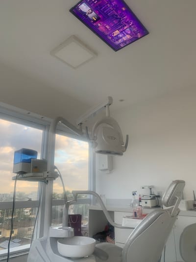 The image depicts an unoccupied, modern dental operatory located in the city of Lauro de Freitas, Brazil. The room features white walls and a white ceiling. Two light fixtures are visible on the ceiling: a square, flush-mounted LED panel and a larger, rectangular display screen exhibiting purple-toned digital art, which includes a partial human profile in the upper left corner.
In the foreground, a grey dental chair is positioned, equipped with a white ceramic spittoon basin and an attached arm supporting a water reservoir with a blue-translucent top. A white dental examination light, designed to illuminate the patient's oral cavity, is suspended overhead and to the right of the chair.
Along the back wall, a white, minimalist countertop holds various dental instruments, including spray bottles, small containers, and a white, table-top appliance, possibly a heating or curing unit. A purple shopping bag is partially visible beneath the counter. A large, floor-to-ceiling window on the left side of the room provides an expansive view of an urban landscape with multiple buildings and trees under a cloudy sky, indicating the operatory is located on an upper floor. No individuals are present in the scene.
