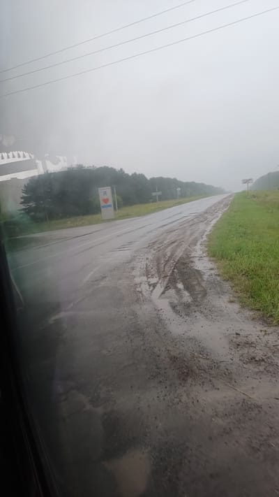 The image shows a wet, rural road in overcast conditions, likely during or after rain. The road surface appears to be a mixture of asphalt and dirt, with visible tire tracks and puddles. On the left side of the road, there is a tall, rectangular sign with a red heart symbol and text that reads "Я Еду" (Ya Yedu), which translates to "I'm going" or "I'm traveling." This sign is likely a landmark or directional marker. Trees and greenery line the sides of the road, suggesting a natural or semi-rural environment. In the background, the road curves and recedes into the hazy distance. The overall atmosphere is muted and possibly a bit somber due to the weather. The context provided indicates this scene is located in the village of Киясово (Kiyasovo), Russia.