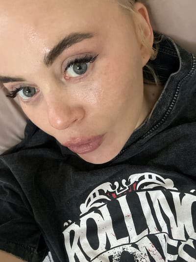 A fair-skinned person with light blonde hair is lying down, as evidenced by the rotated perspective. The person's face is prominent, with focus on their eyes, nose, and lips. They have dark eyebrows and long eyelashes. They are wearing a dark-colored shirt with a white graphic design. The background is a light, solid color, possibly a bed or similar soft surface. There's no information available in the image to associate it with the city of Высокий, Russia.