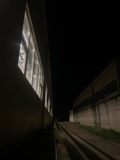 The image depicts an industrial area at night in Kazanlak, Bulgaria. On the left, a light-colored building with a row of illuminated windows runs parallel to a dark path. The windows are rectangular and appear to be part of an office or factory. To the right of the path is a concrete building with a long, dark band of windows along its upper section. The path itself is unpaved and shows distinct shadows cast by unseen objects, suggesting artificial lighting. Patches of grass grow alongside the path and against the concrete building. The overall scene is dark and somewhat desolate, characteristic of an industrial zone after business hours. There are no people or animals visible in the image.