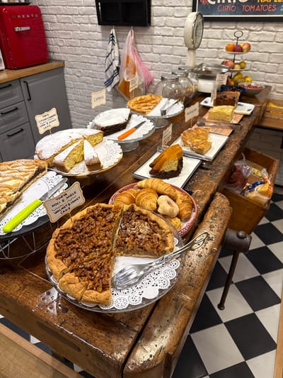 This media file shows a close-up, overhead view of a display of baked goods, likely in a bakery or cafe in Naples, Italy. The focus is on an assortment of cakes, tarts, and pastries arranged on a rustic wooden counter. The display includes slices of what appear to be pound cake dusted with powdered sugar, a chocolate and orange layered cake, a large pecan or walnut tart, and croissants and donuts in a pink woven basket. Several small labels with text written in Italian identify some of the items, such as "CAPRESE", "TORTA LIMONE E MANDORLE", and "TORTA NOCI E MIELE".
In the background, a black and white checkered tile pattern is visible on the wall, and a vintage-style metal scale with a white dial sits to the left. A sign with text that includes "CIRIO TOMATOES" is also present. The lighting suggests it might be daytime, possibly with artificial light contributing to the scene. The overall impression is one of a well-stocked bakery counter, inviting customers to choose from a variety of sweet treats. The rustic wooden counter and the vintage scale add to a charming, perhaps traditional, atmosphere.
