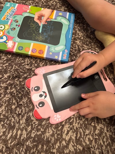Two children's drawing tablets are on a brown carpet. The tablet on the left is green and shaped like a cat. It is inside its original packaging, which is colorful and features a drawing of a hand using a stylus. The tablet on the right is pink and shaped like a bear. A child's hand is holding a black stylus and drawing on the pink tablet. The location is not specified in the image.