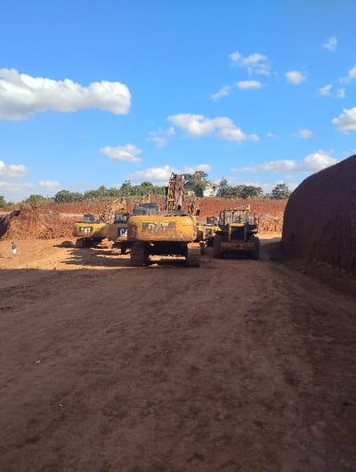 This image depicts an active excavation site in Kikuyu, Kenya, under a clear blue sky with scattered white cumulus clouds. The ground consists of reddish-brown disturbed earth. In the midground, a cluster of yellow heavy construction vehicles is stationary. The most prominent vehicle is a large, tracked excavator, viewed from the rear, bearing "CAT" branding, positioned centrally. To its left and slightly behind are additional yellow CAT excavators, one with its boom partially raised. To the right of the central excavator, a yellow wheeled loader is partially visible. The background features excavated earth banks and mounds of reddish-brown soil. Beyond the excavation zone, a line of green vegetation is visible, with distant light-colored buildings partially observable above the tree line. A substantial, rounded mound of reddish-brown earth occupies the right side of the frame, rising high. A single individual, dressed in light-colored clothing, stands on the ground in the far left midground. No active work or interactions are observed.