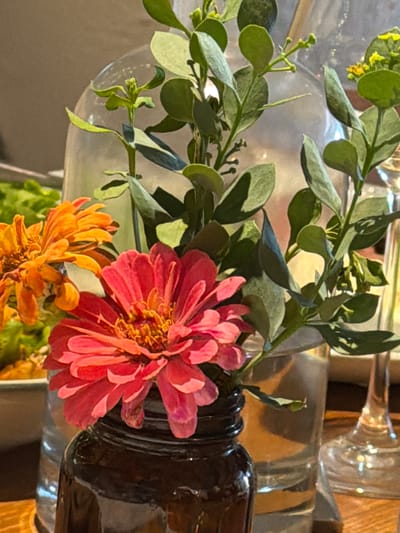 This close-up image captures a vibrant floral arrangement featuring two prominent zinnia-like flowers amidst green foliage. In the foreground, a large, deeply-hued pink to red flower with multiple layers of petals and a yellow-orange center dominates the right side. Below and slightly to its left, another flower, similar in structure but in shades of orange and yellow, is partially visible.
The flowers are arranged with several sprigs of green leafy branches, possibly eucalyptus or a similar decorative plant, which extend across the upper and left portions of the frame. The leaves are generally oval or rounded, and some branches show small buds or developing clusters of greenery.
The arrangement is held in various glass and dark-colored containers, which are partially obscured and largely out of focus. A dark, possibly brown, ribbed bottle or vase is visible on the right, providing a dark contrast to the bright flowers. Other clear glass vessels are seen in the background, reflecting light and creating soft, blurry highlights. The overall setting appears to be indoors, bathed in natural light, suggesting it is daytime. There are no people visible, and no text or specific identifying location cues are present within the image itself. The scene is presented as a still life, focusing on the beauty and color of the flowers and greenery. The image evokes a sense of freshness and warmth.