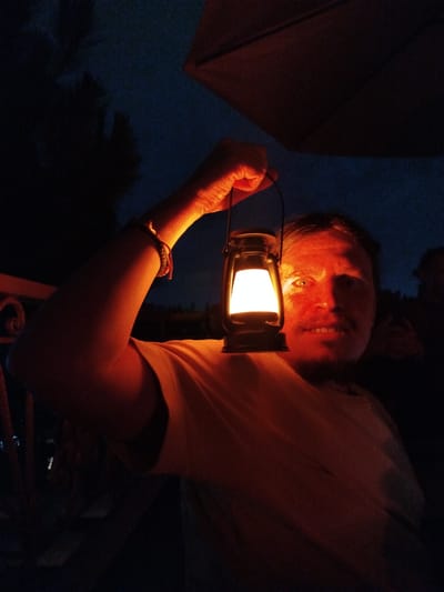 A man holds a lit lantern aloft in a dark setting. The man is fair-skinned and wears a light-colored T-shirt. His face is illuminated by the lantern's glow, casting strong shadows. He has a bracelet on his raised arm. The scene appears to be outdoors, with suggestions of foliage and an umbrella-like structure in the background. The location is undetermined.