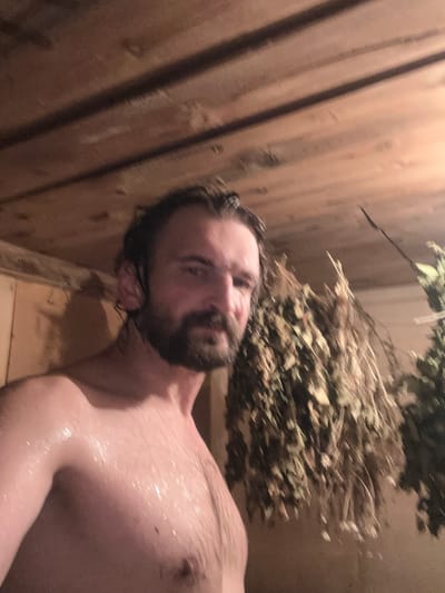 The image shows a man in a wooden sauna, likely in Biysk, Russia. He is partially undressed, with sweat visible on his skin. The walls and ceiling of the sauna are made of wood. Bunches of dried leaves, possibly for a banya (traditional Russian bathhouse), hang from the ceiling. The man has a beard and wet hair, and he is looking at the camera.