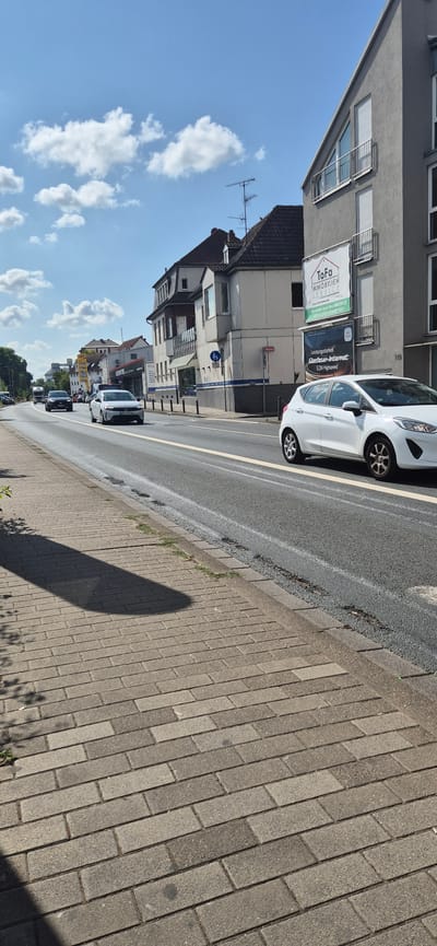 This is a slightly low-angle, full-shot photograph taken on a bright, sunny day in Mainz, Germany, on Friedrich-Ebert-Straße. The image captures a street scene looking down the road. On the right side of the image, a modern, gray, multi-story building is prominent, featuring multiple windows with white shutters. A large white banner advertising "ToFa Immobilien Service" and "Leistungsstarkes Glasfaser-Internet" is affixed to its facade. A white Ford Fiesta is driving on the asphalt road in front of this building. The road has a clear white line marking its center and a yellow line on the right edge. Cars are visible in the distance, traveling in both directions. On the left side of the road, a sidewalk paved with gray bricks is visible, with a long shadow stretching across it from an unseen object to the left. Beyond the sidewalk, a line of trees and more buildings with pitched roofs can be seen, some with older architectural styles. The sky is a vibrant blue with scattered white clouds.