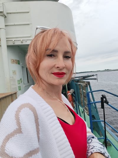 A woman with short, peachy-pink hair and bright red lipstick takes a selfie on the deck of a ferry. She is wearing a white cardigan with a large, light brown heart design on the left side and a red tank top underneath. Her head is slightly tilted, and she is smiling faintly. Behind her, the ferry railing, a life preserver, and the calm, grey water of a river or lake are visible. The sky is overcast, suggesting a cloudy day. The setting appears to be a waterway in Russia, given the "Bereg, Russia" location context. There are some signs with symbols on a white wall behind her, but the text is not legible. The overall mood is casual and relaxed, with the woman enjoying her time on the water.