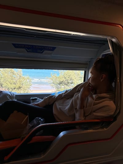 A person is looking out a window of a vehicle, possibly a bus or train, with a view of a beach and trees. The person is wearing a light-colored hoodie and dark pants. They are positioned inside the vehicle, which appears to be a limosine bus, as indicated by the text "NAM HAI LIMOUSINE" visible on a blue sticker on the interior wall. The lighting suggests it could be daytime, with natural light coming in through the window. The overall mood appears contemplative or curious as the person gazes outwards. The scene outside the window shows a bright, sunny day with a calm body of water and a sandy beach, bordered by lush green trees. The interior of the vehicle shows some luggage or personal belongings stored near the person. The image is taken from a slightly low angle, looking upwards towards the person and the window.