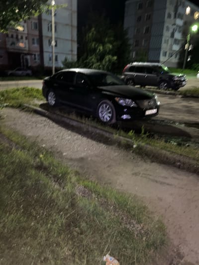 An exterior, eye-level, nighttime, slightly blurry photograph shows a black sedan parked on a dirt path beside a grassy embankment. The sedan is a Lexus LS460, identified by its grill and headlights. Its license plate is partially obscured but reads "У616ХУ". The car is parked in front of an apartment building in the city of Kokhma, Russia. Another dark-colored SUV is parked behind the sedan, also on the dirt path. The environment is residential, with tall apartment buildings visible in the background. The lighting is artificial, likely from streetlights, casting shadows and creating a slightly eerie atmosphere. The overall scene depicts a common urban parking situation at night.