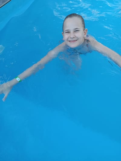 A young girl with blonde hair pulled back smiles at the camera while swimming in a bright blue pool. She is positioned in the center of the frame, with her head and upper body visible. Her arms are extended to her sides, creating ripples in the water. A green and white wristband is visible on her left arm. The water appears clear and inviting, suggesting a warm, sunny day suitable for swimming. The overall mood is cheerful and playful, characteristic of a recreational activity. The background is entirely composed of the blue water, with some subtle light reflections and disturbances on the surface indicating movement. There is no visible text in the image.