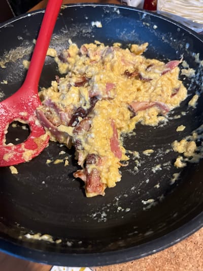 An overhead shot captures scrambled eggs with bacon and feta cheese in a black frying pan. The eggs appear creamy yellow with distinct curds, intermingled with pieces of bacon or ham. Some meat pieces are dark and crispy, while others are lighter and softer. White crumbles, consistent with feta cheese, are visibly mixed throughout the egg dish.
A red silicone spatula rests in the lower left portion of the pan, partially covered in the egg mixture. The pan itself is dark and shows some residue on its interior sides, suggesting the food has just been cooked or is being stirred. The activity depicted is the preparation or immediate aftermath of cooking scrambled eggs with additions.
In the background, subtle hints of a cork-like surface are visible on the right edge, and a light-colored object, possibly paper or a napkin, can be seen on the far left. There is no visible text in the image. The scene conveys a home cooking environment, likely for a breakfast or brunch meal, within the provided location context of Chêne-Bourg, Switzerland.