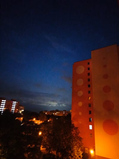 This is a night-time, low-light photograph of an urban environment in the city of Most, Czechia. The scene is dominated by a deep blue sky with some faint clouds and distant city lights.
On the right side of the image, a tall, orange-colored apartment building is prominently featured. This building has a distinctive design with large, circular, lighter-colored shapes and several dark, rectangular windows. One window on the rightmost building emits a bright white light.
To the left of the orange building, further in the background, is another apartment building, also illuminated with many windows showing light.
In the foreground and middle ground, the tops of trees are visible, silhouetted against the urban lights and the twilight sky. Several streetlights cast a warm, yellow glow on the trees and surrounding area, indicating a residential or commercial street below. The overall impression is one of a quiet, residential area at dusk or after dark.