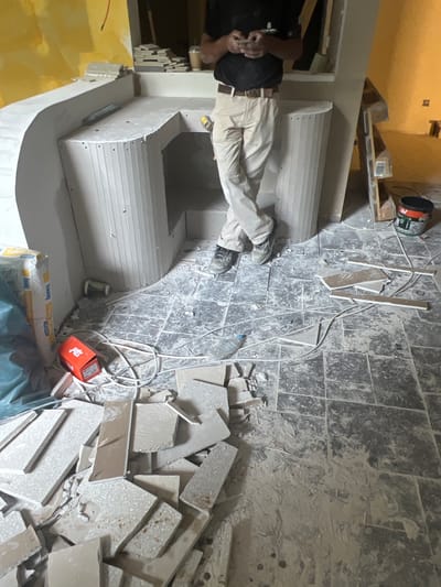 The image depicts an indoor construction or renovation site, possibly a kitchen or bathroom, in Athens, Greece. A man, wearing a black t-shirt, light-colored trousers, and dark shoes, stands with his legs crossed, holding a mobile phone. He is positioned in front of a white, unfinished counter structure that appears to be made of drywall or plaster. The floor is tiled with dark, square tiles, and is covered in dust and debris, including broken tiles and construction materials. Various construction supplies are scattered around the area, such as bags of plaster, rolls of wire, and a red box. In the background, part of a yellow-painted wall is visible. The overall scene suggests an ongoing project with a focus on the installation or finishing of the counter and floor.
