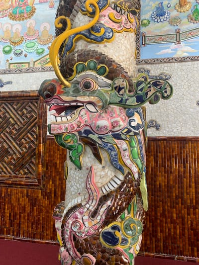 In Xuân Thành, Vietnam, a close-up shot focuses on a ceramic dragon pillar in a temple. The dragon's head, adorned with intricate mosaic pieces, is prominently displayed, with its mouth open to reveal a row of teeth. The pillar itself is covered in a mosaic of small, white ceramic tiles, creating a textured surface. Behind the pillar, a wall is decorated with two distinct patterns. The upper section features a mural with a pale blue sky, clouds, and figures of people in prayer. Below this mural, the wall is covered in vertical, reddish-brown ceramic tiles, arranged in a brick-like pattern. To the left of the pillar, a section of the wall is covered with a brown, lattice-like decorative panel, also made of ceramic tiles. The floor is covered with a dark red carpet. The lighting is bright, casting subtle shadows and highlighting the vibrant colors and detailed craftsmanship of the dragon pillar and the surrounding temple decor.