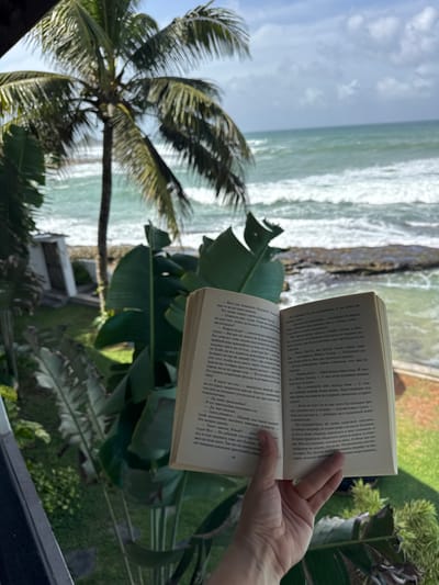 An open book is held by a person's hand in the foreground, with a lush green palm tree and the ocean in the background. The book is open to pages with text. The scene appears to be outdoors, possibly on a balcony or patio, overlooking a tropical beach. The lighting suggests it is daytime. The location is identified as Goviyapana, Sri Lanka.