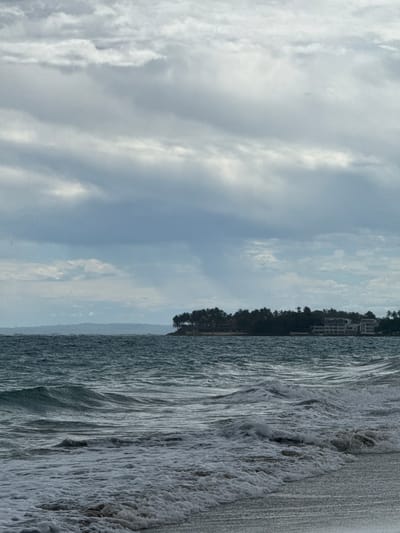 The image depicts a vertical view of a shoreline in Cabarete, Dominican Republic. The sky is cloudy, with the shoreline showing a line of trees, some buildings. The ocean is active with waves crashing near the sandy beach.