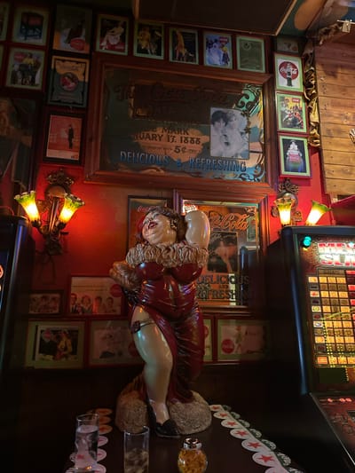 This is a vertically oriented image of a room, likely a bar or a diner, decorated with vintage Coca-Cola memorabilia and other assorted items. The overall scene is dimly lit, with warm lighting emanating from two wall sconces featuring green glass shades and from a slot machine in the background.
Dominating the central portion of the image is a large, life-sized statue of a voluptuous woman in a red dress, posed provocatively. She appears to be made of plastic or resin and is positioned on a textured base.
The walls are adorned with numerous framed items, predominantly vintage Coca-Cola advertisements and posters. These range from individual bottle depictions to larger, more elaborate designs featuring text like "DELICIOUS & REFRESHING" and dates such as "JANUARY 13. 1888". Some frames are dark wood, while others have a metallic or more ornate appearance.
In the upper right, a slot machine is partially visible, with glowing lights and rows of symbols, suggesting an entertainment or gaming element to the venue. Along the right edge of the image, a series of Heineken beer coasters are strung together, further contributing to the bar-like atmosphere.
Other objects include what appears to be a speaker system on the left, and a glass with snacks or chips on the far right. The background is composed of wooden planks, adding to a rustic or old-fashioned aesthetic. The presence of Dutch text on some of the advertisements ("TRACTATIE VOOR ALLE VIER", "KENMERK VAY") confirms the location as the Netherlands. The overall mood is eclectic and retro, with a focus on nostalgic branding and entertainment.